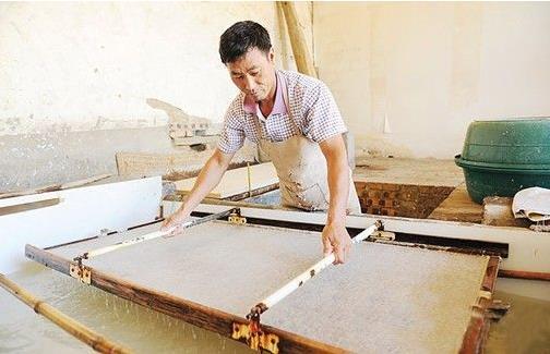 圖中的造紙師傅手執竹簾，嫻熟地在紙漿池中撈起濕紙。